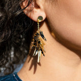 Close-up of a person wearing featherlight, gold dangle earrings with multiple shiny, leaf-like pieces, set against curly hair and denim clothing.
