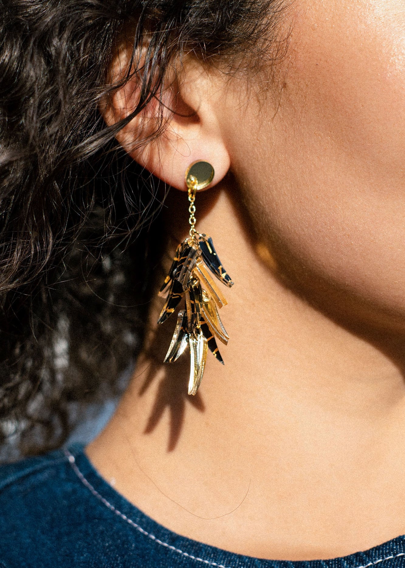 Close-up of a person wearing featherlight, gold dangle earrings with multiple shiny, leaf-like pieces, set against curly hair and denim clothing.