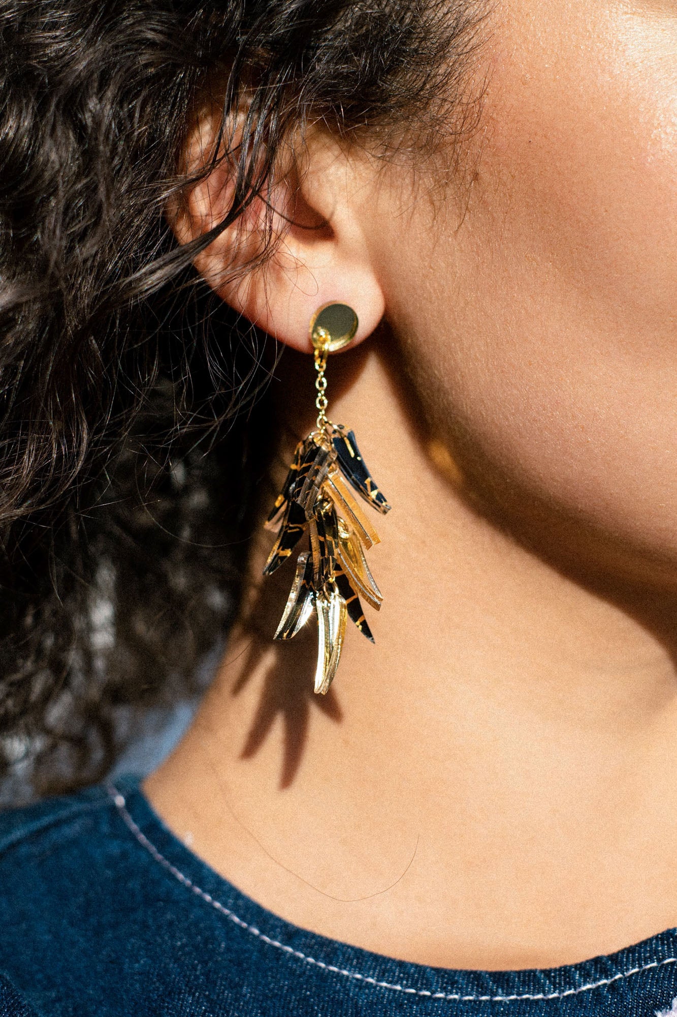 Close-up of a person wearing featherlight, gold dangle earrings with multiple shiny, leaf-like pieces, set against curly hair and denim clothing.