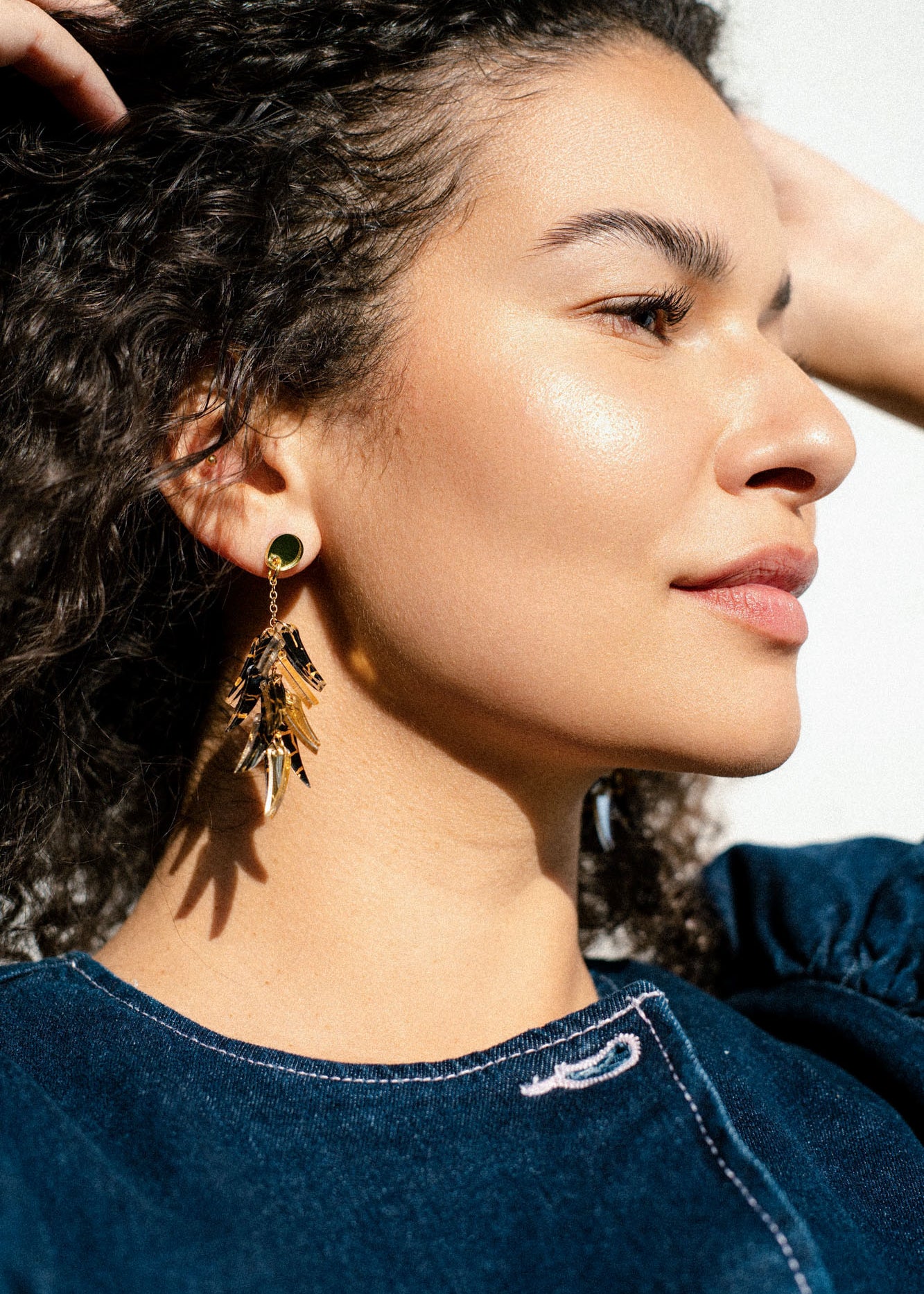 A woman with curly hair wearing a blue top and featherlight gold dangle earrings is shown in close-up, looking to the side in bright, natural light.