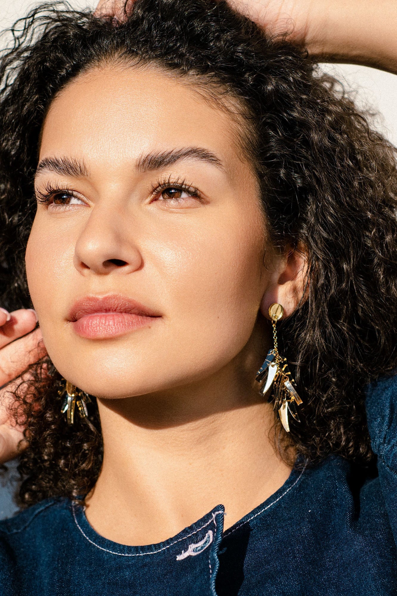 A woman with curly hair and glowing skin gazes into the distance, wearing featherlight gold dangle earrings and a dark blue top. Sunlight highlights her features as she gently touches her hair with one hand.