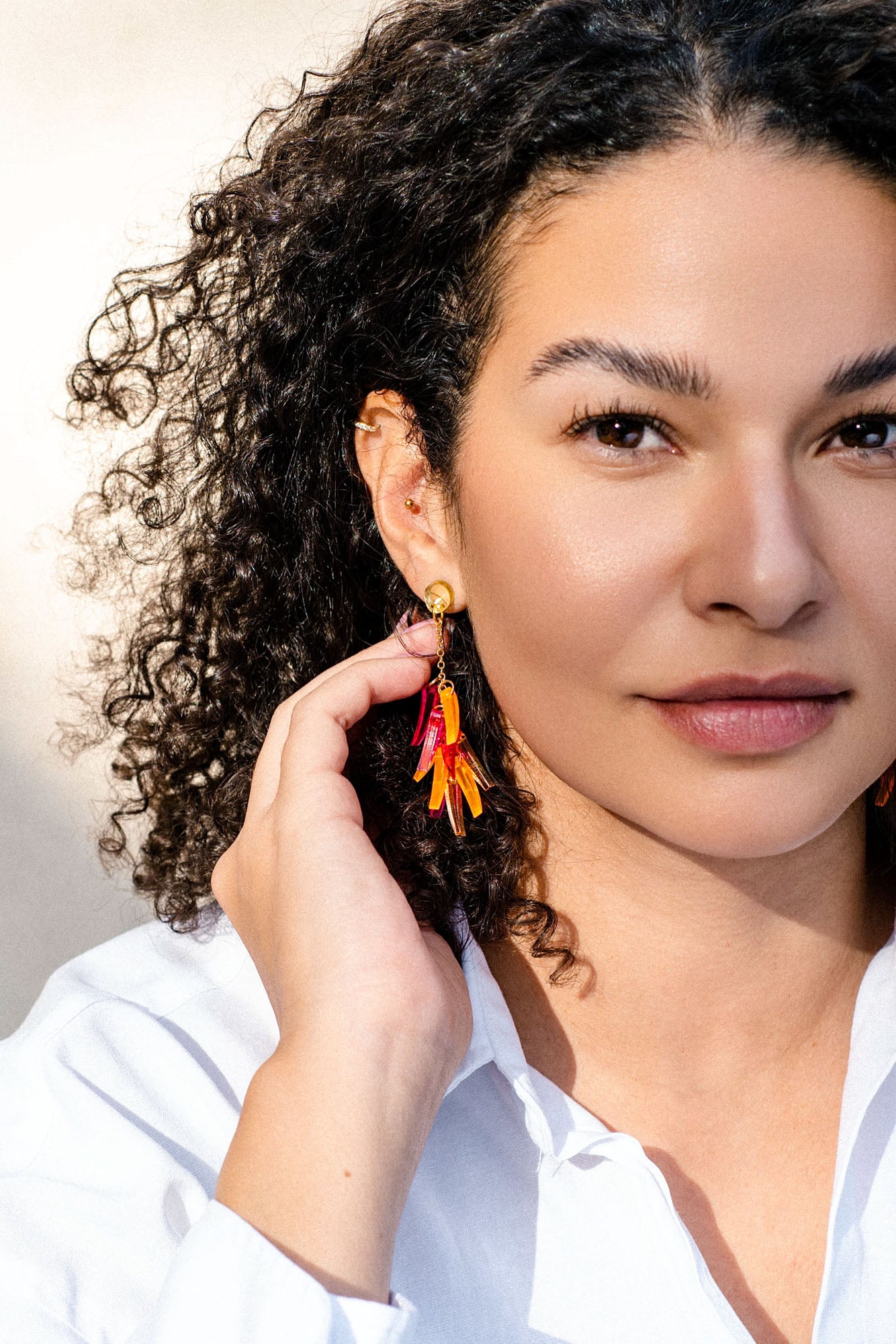 A woman with curly hair in a white shirt holds her hand to her ear, showing off featherlight orange and red dangle earrings. She has a calm expression and natural makeup against a bright, neutral background.