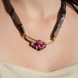 A woman wearing a cream-colored top and a unique handmade necklace featuring a fabric band, gold accents, and a cluster of pink sculpted beads as the centerpiece.