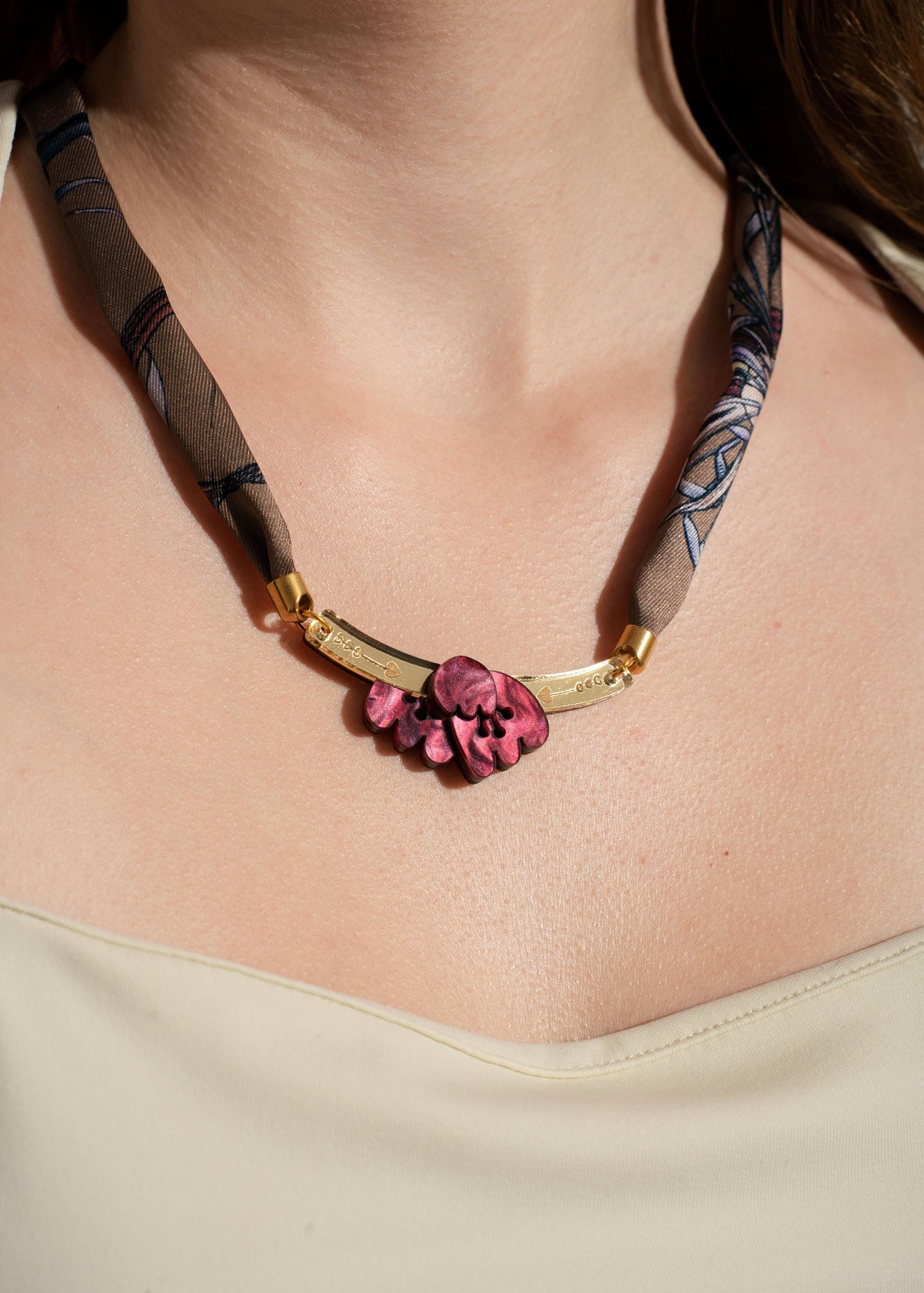 A woman wearing a cream-colored top and a unique handmade necklace featuring a fabric band, gold accents, and a cluster of pink sculpted beads as the centerpiece.