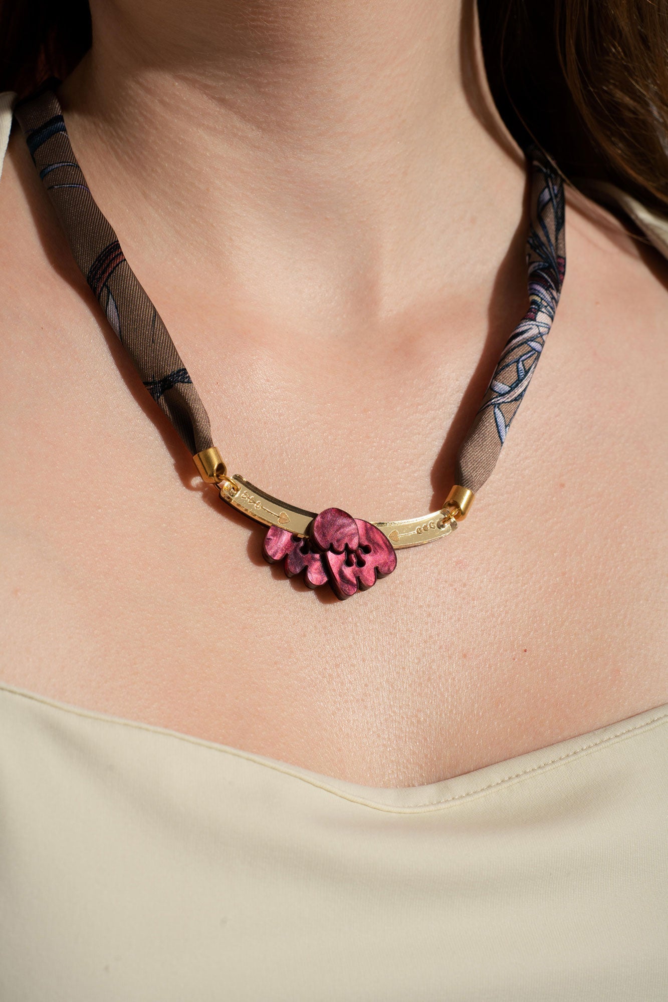 A woman wearing a cream-colored top and a unique handmade necklace featuring a fabric band, gold accents, and a cluster of pink sculpted beads as the centerpiece.
