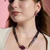 Woman with light brown hair wearing a cream top, maroon floral earrings, and a matching handmade necklace, standing against a pink background.