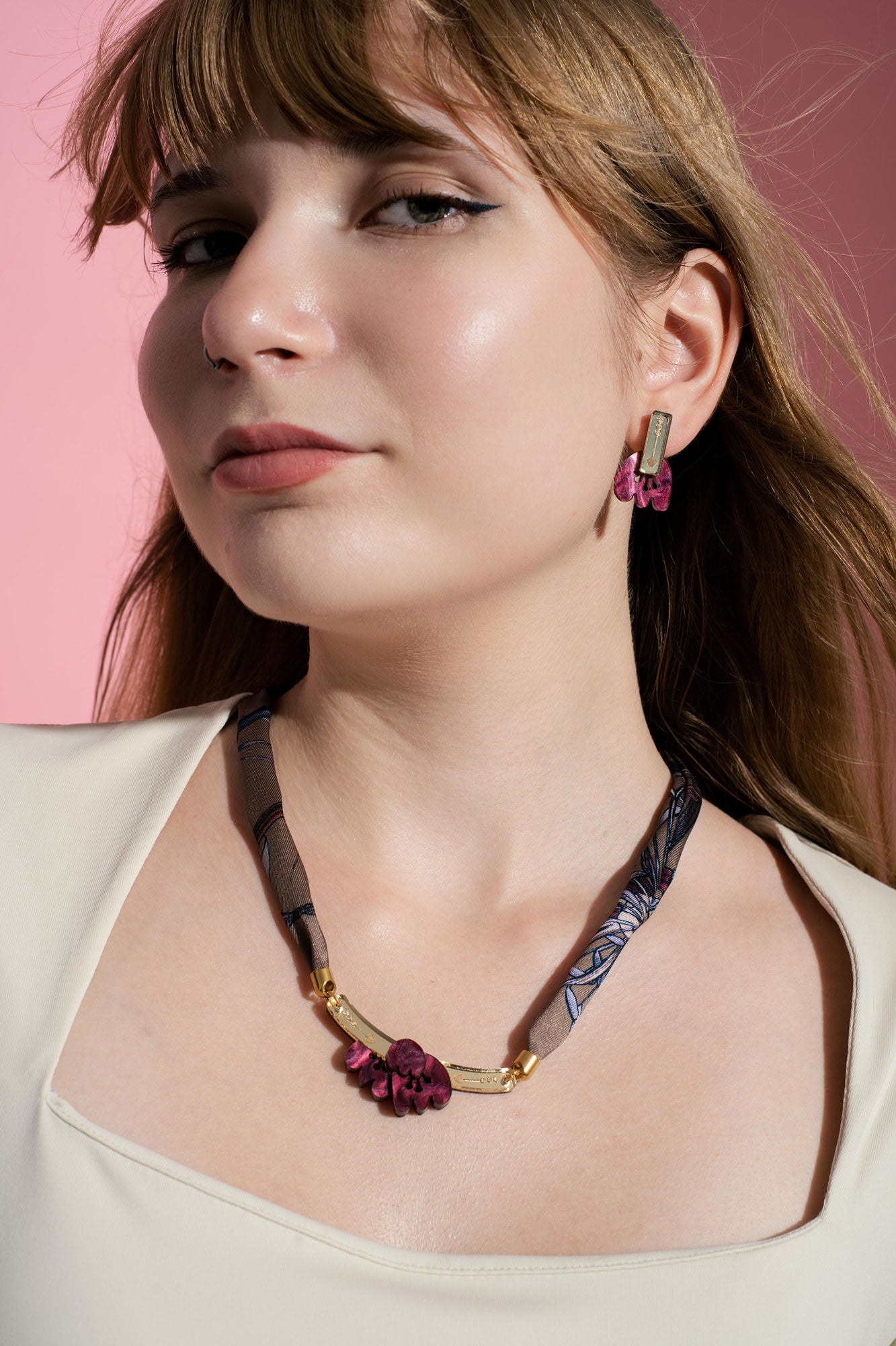Woman with light brown hair wearing a cream top, maroon floral earrings, and a matching handmade necklace, standing against a pink background.