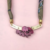 A handmade necklace with a fabric strap, gold bar, and pink flower pendant is displayed on a pink background. The bar is decorated with small heart and dot engravings, while the design evokes the charm of an acrylic tulip paired with the elegance of a silk-print scarf.