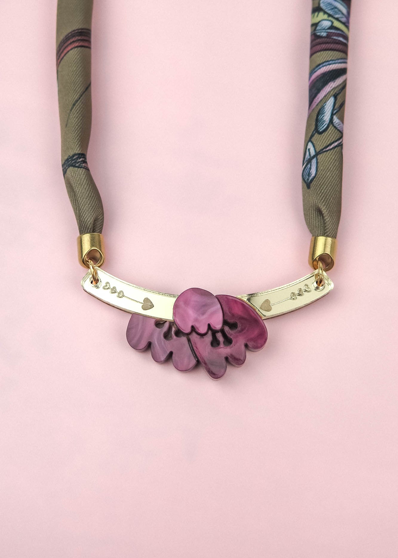 A handmade necklace with a fabric strap, gold bar, and pink flower pendant is displayed on a pink background. The bar is decorated with small heart and dot engravings, while the design evokes the charm of an acrylic tulip paired with the elegance of a silk-print scarf.