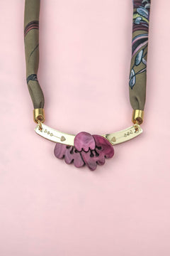 A handmade necklace with a fabric strap, gold bar, and pink flower pendant is displayed on a pink background. The bar is decorated with small heart and dot engravings, while the design evokes the charm of an acrylic tulip paired with the elegance of a silk-print scarf.