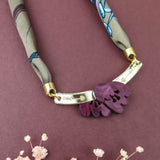 A fabric and metal handmade necklace with purple leaf-shaped charms on a maroon background, accented by small dried flowers at the bottom—perfect to pair with a silk-print scarf or your favorite acrylic tulip accessories.