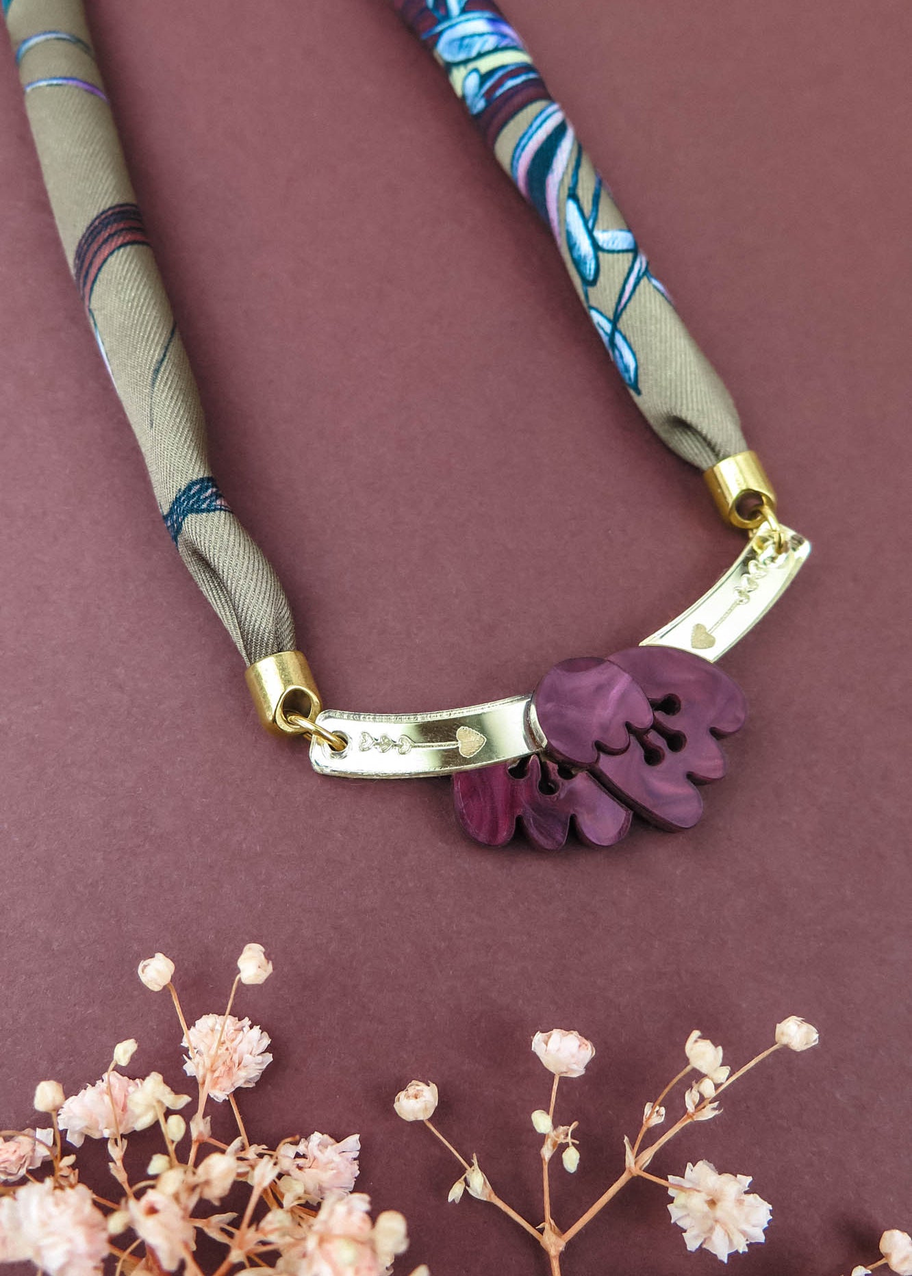A fabric and metal handmade necklace with purple leaf-shaped charms on a maroon background, accented by small dried flowers at the bottom—perfect to pair with a silk-print scarf or your favorite acrylic tulip accessories.