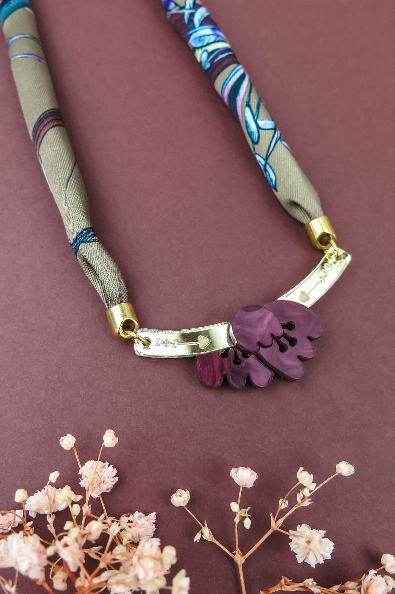 A fabric and metal handmade necklace with purple leaf-shaped charms on a maroon background, accented by small dried flowers at the bottom—perfect to pair with a silk-print scarf or your favorite acrylic tulip accessories.