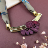 A handmade necklace featuring a purple floral pendant with gold details and a patterned fabric strap, displayed on a mauve surface with small pink dried flowers and a cream silk-print scarf in the background.