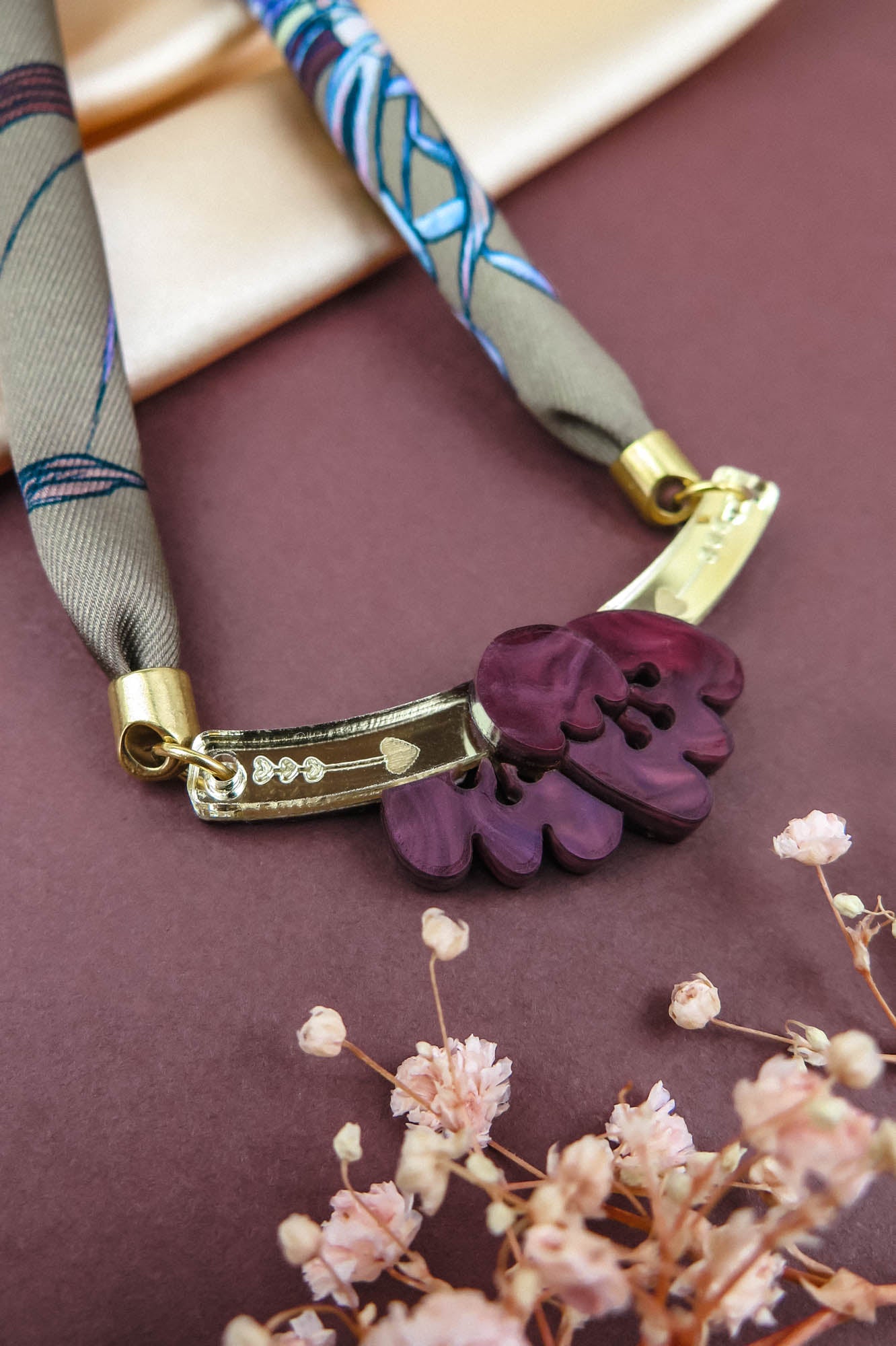 A handmade necklace featuring a purple floral pendant with gold details and a patterned fabric strap, displayed on a mauve surface with small pink dried flowers and a cream silk-print scarf in the background.