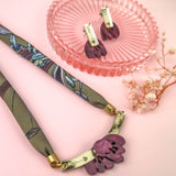 A handmade necklace with a floral pendant and matching earrings, both featuring purple acrylic tulip charms, are displayed on a pink surface with a pink glass dish and a small dried flower.