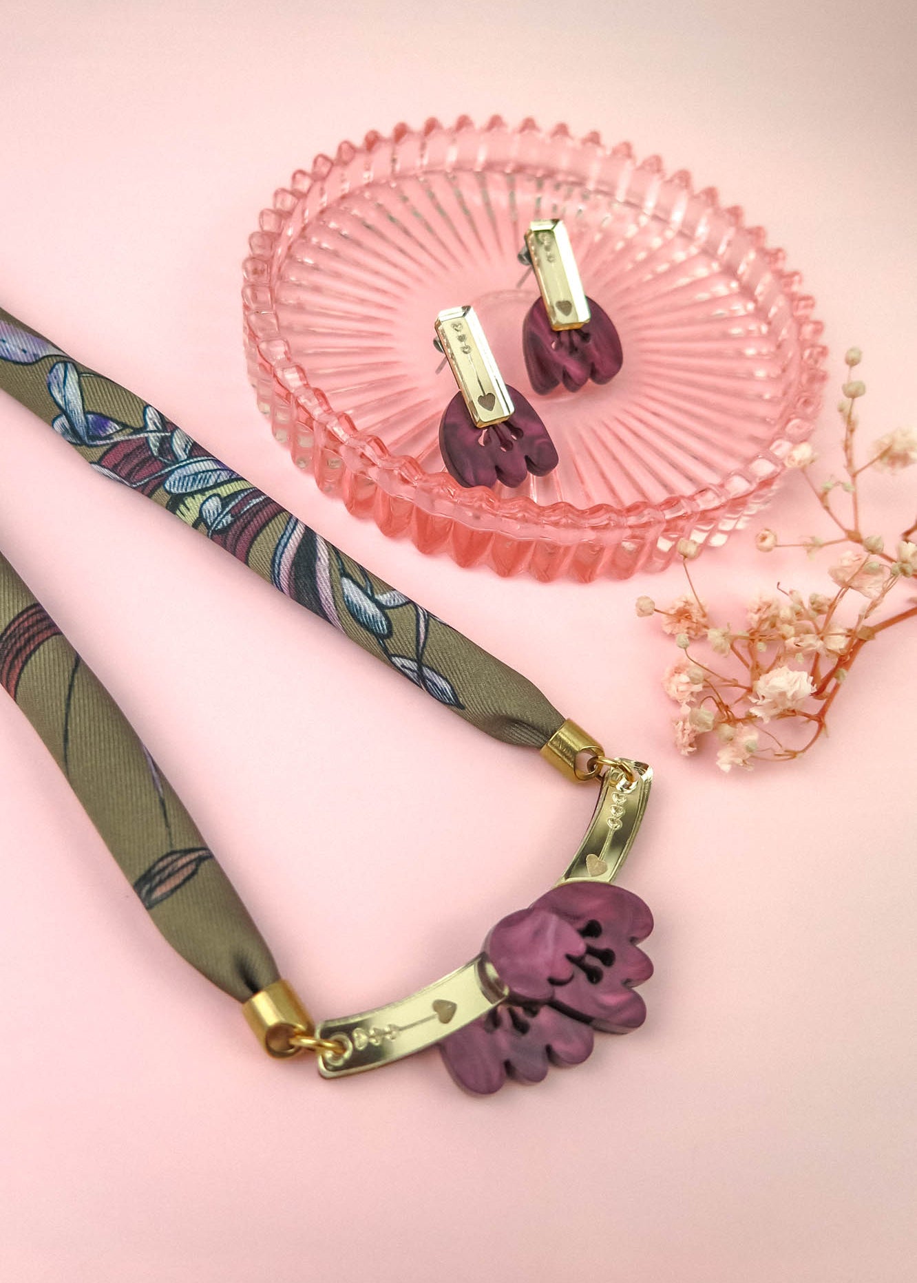 A handmade necklace with a floral pendant and matching earrings, both featuring purple acrylic tulip charms, are displayed on a pink surface with a pink glass dish and a small dried flower.