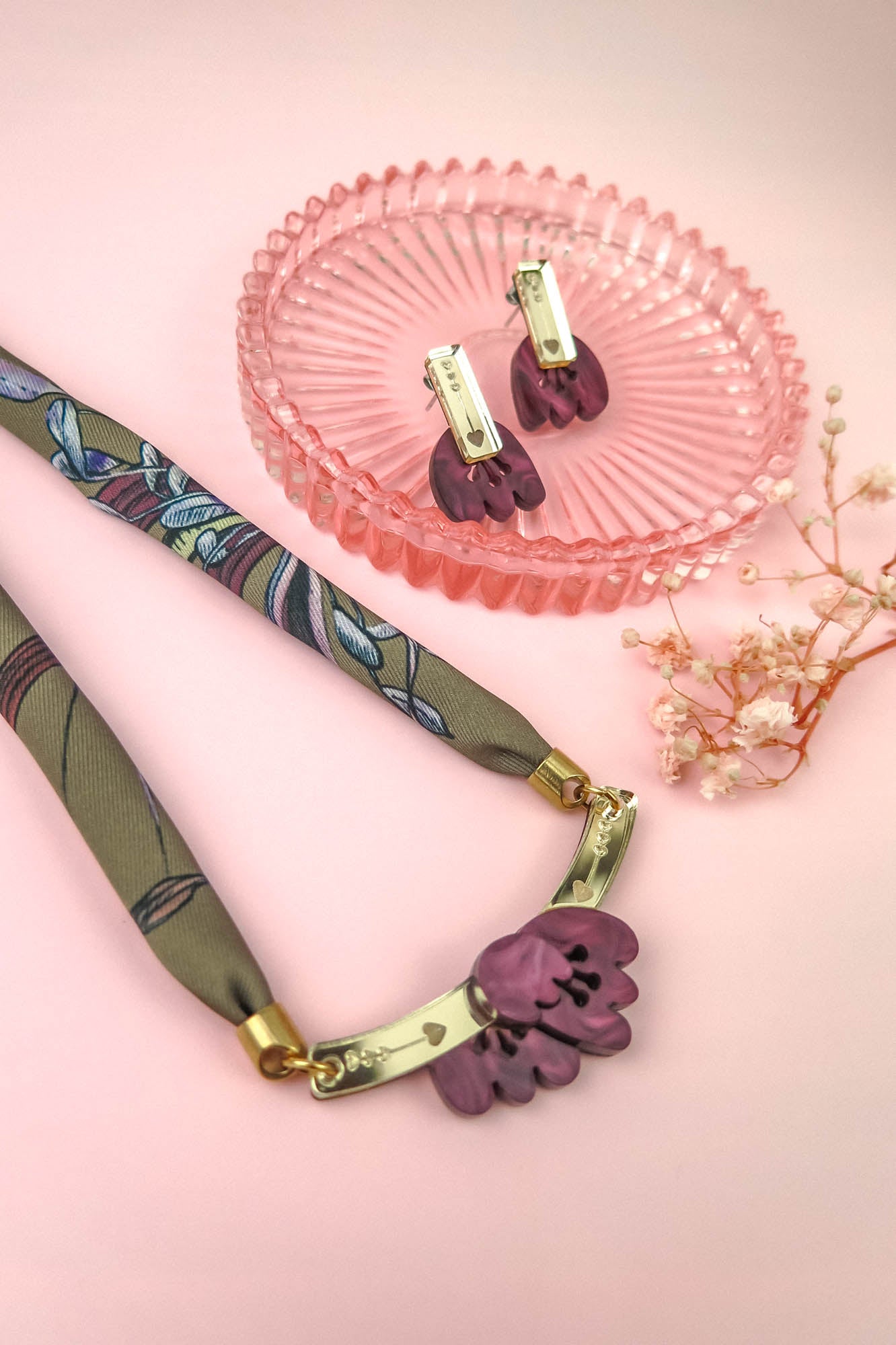 A handmade necklace with a floral pendant and matching earrings, both featuring purple acrylic tulip charms, are displayed on a pink surface with a pink glass dish and a small dried flower.