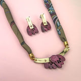 A necklace and matching earrings with purple floral pendants, gold accents, and a patterned fabric cord are displayed on a pink background, featuring delicate acrylic tulip details for a charming touch.