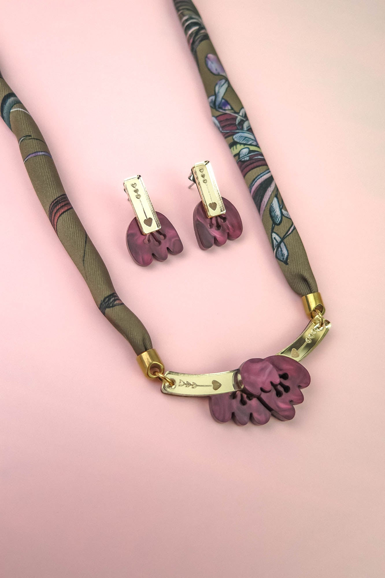 A necklace and matching earrings with purple floral pendants, gold accents, and a patterned fabric cord are displayed on a pink background, featuring delicate acrylic tulip details for a charming touch.