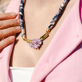A person wearing a pink shirt and white top displays a handmade necklace with a patterned fabric band and gold clasp, featuring pink and purple gemstone flowers. Their hand rests near the collarbone, complementing an elegant silk-print scarf.