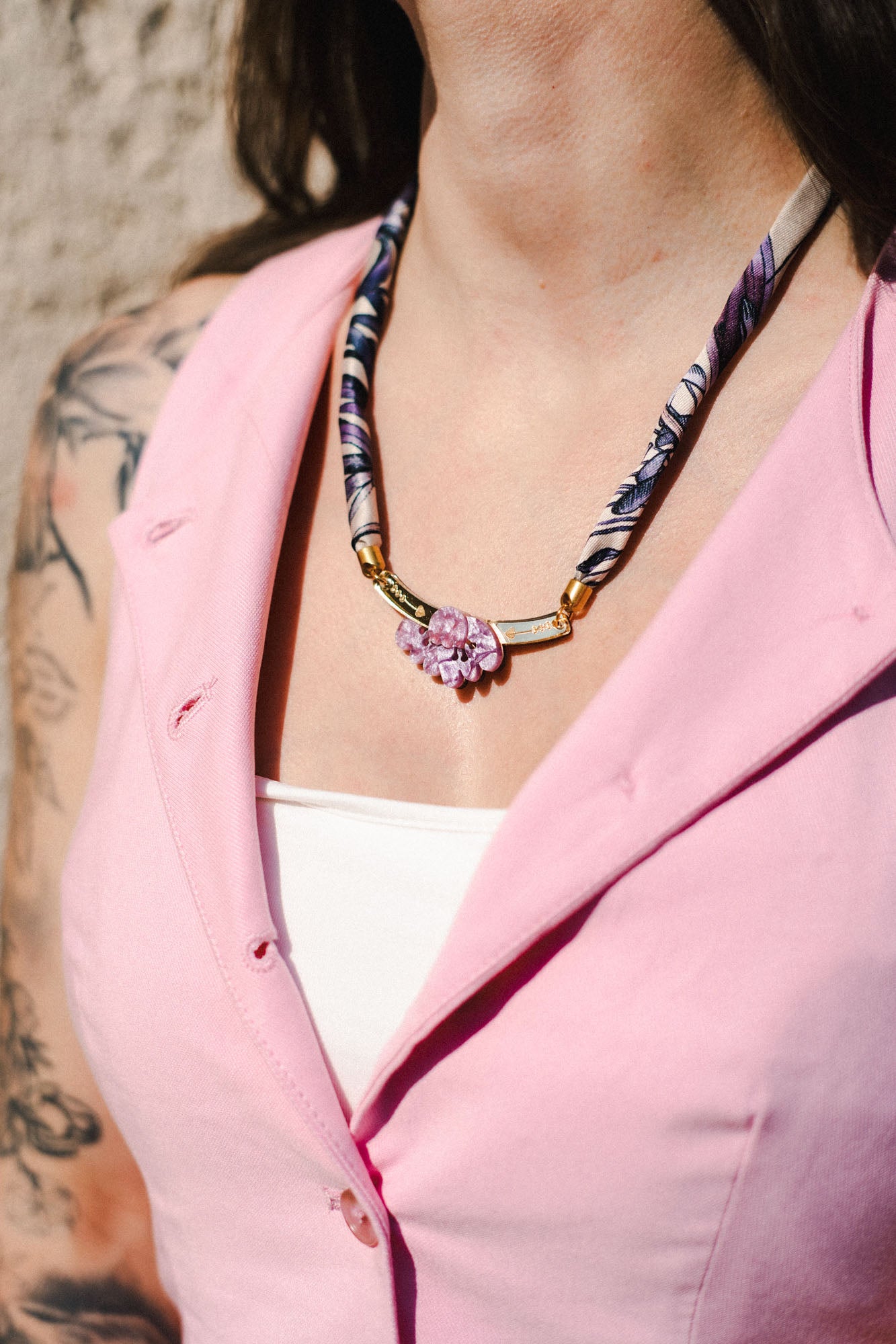 A woman wearing a light pink vest over a white top showcases a handmade necklace with a pink flower pendant; her arm features visible tattoos.