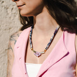 A woman with long dark hair and a floral tattoo on her arm wears a pink sleeveless vest, a white top, and a handmade necklace with a pink pendant, standing outdoors in sunlight.