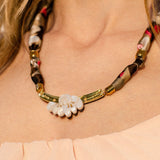 A close-up of a woman wearing a handmade necklace featuring an acrylic tulip pendant, gold accents, and an upcycled fabric-wrapped chain in brown, beige, and red tones; she is dressed in a peach off-shoulder top.