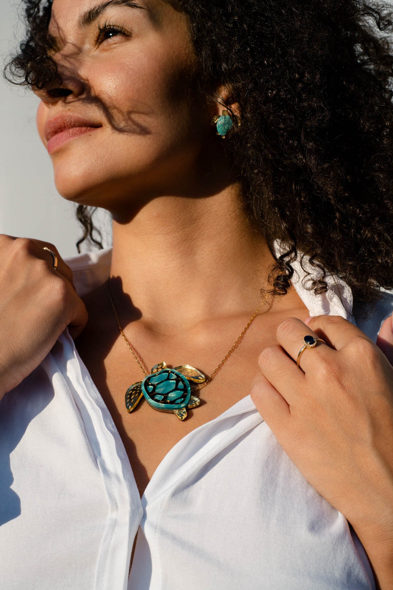 A woman in a white shirt wears a stunning turquoise and gold Sea Turtle Necklace, complemented by turquoise earrings and rings. Sunlight highlights her face and curly hair as she looks upward, casting a shadow across her cheek—a perfect moment to showcase her love for handcrafted jewelry.
